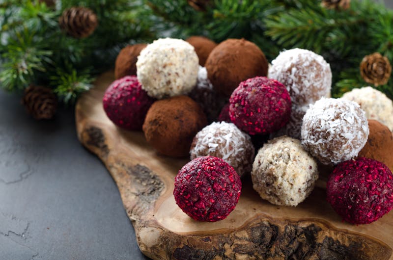 Homemade vegan chocolate candies with dried raspberry, grated coconut, nuts, cocoa on a wooden board made of olive tree and dark stone background. Fir branches on the back. Selective focus, copyspace
