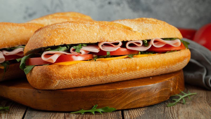 Two Long baguette Sandwiches with arugula, slices of fresh tomatoes, ham and cheese