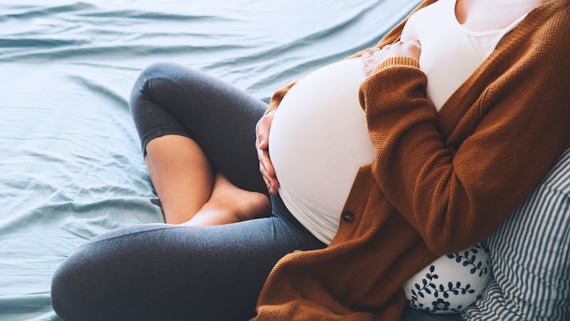 Beautiful pregnant woman sitting at bed and holds hands on belly in bedroom at home.