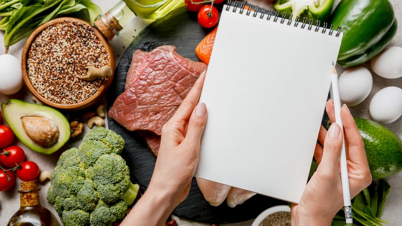 cropped view of woman holding empty notebook above food for ketogenic diet menu