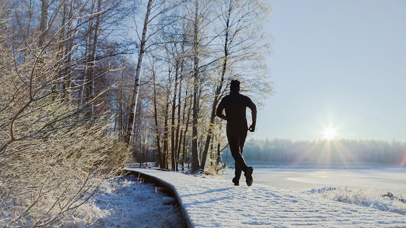 Young, adult alone man in tracksuit and hat running on forest trail in sunny winter morning after first snow. Enjoying sport in sunrise. Daily active lifestyle. Life motion. Back view.