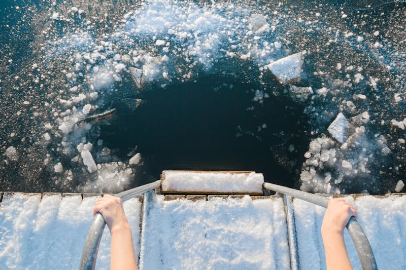 Young woman’s hands holding on metallic ladder handles. Ready for swimming in ice hole at lake after sauna in cold day. Care about body health in winter time. Point of view shot. Close up.