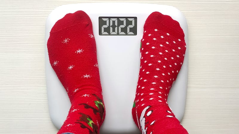 Female feet standing on electronic scales for weight control in funny red Christmas socks on white wooden background.