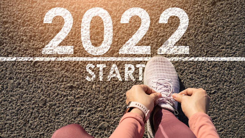 New year 2022 or start straight concept.word 2022 written on the asphalt road and athlete woman runner stretching leg preparing for new year at sunset.Concept of challenge or career path and change.