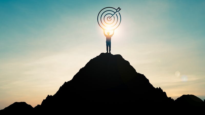 Silhouette of businessman holding target board on the top of mountain with over blue sky and sunlight. It is symbol of leadership successful achievement with goal and objective target.