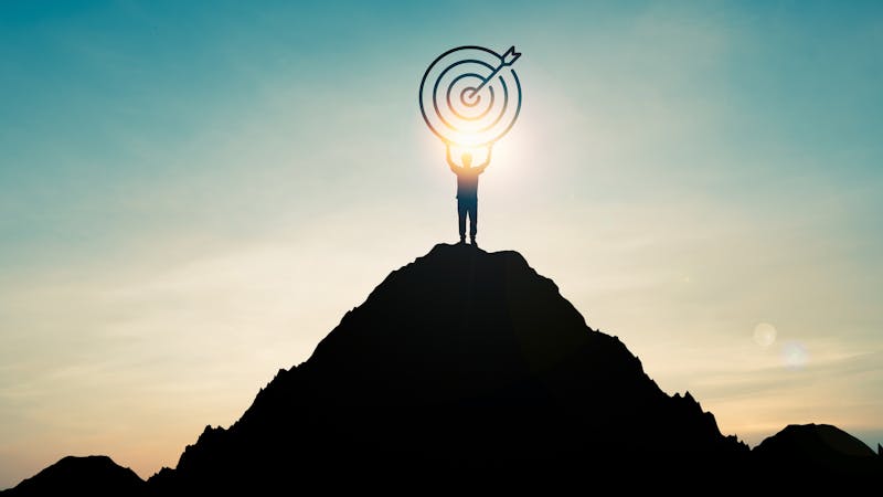 Silhouette of businessman holding target board on the top of mountain with over blue sky and sunlight. It is symbol of leadership successful achievement with goal and objective target.