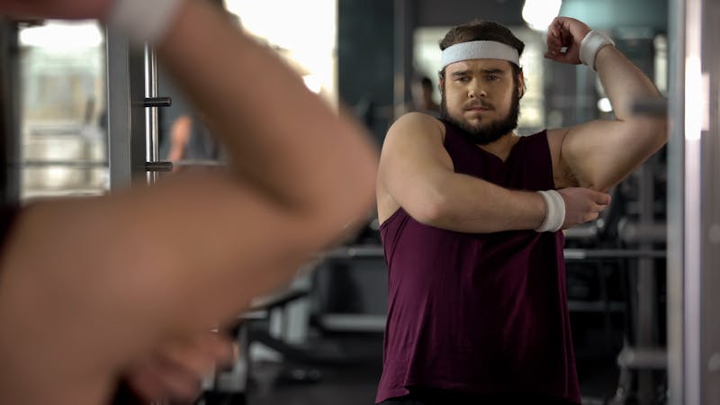 Frustrated overweight man looking at his hand fat dreaming about biceps, obesity