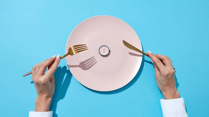 White plate with round whatch shows six o’clock served knife and fork in a girl’s hands on a blue background. Time to eat and diet concept. Top view.