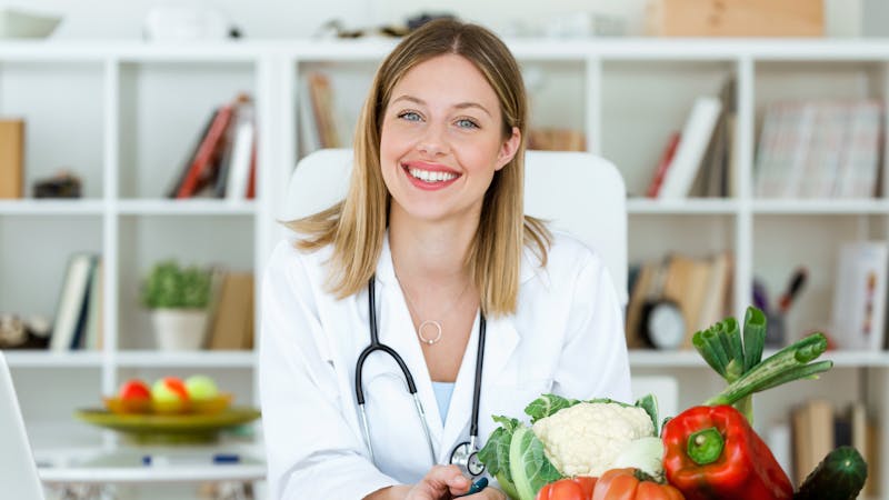 Beautiful smiling nutritionist looking at camera and showing healthy vegetables in the consultation.