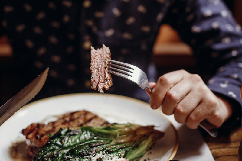 Hands holding fork and knife and eating delicious juicy steak with grilled cabbage,tomatoes and cheese on table at cafe in city street. Man tasting bbq with vegetables in restaurant