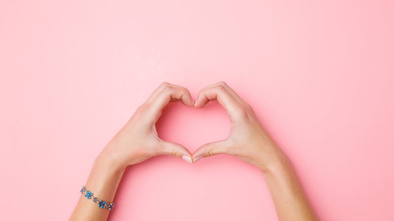 Heart shape created from young woman’s hands on pastel pink background. Love and happiness concept. Empty place for emotional, sentimental text, quote or sayings. Closeup. Top view.