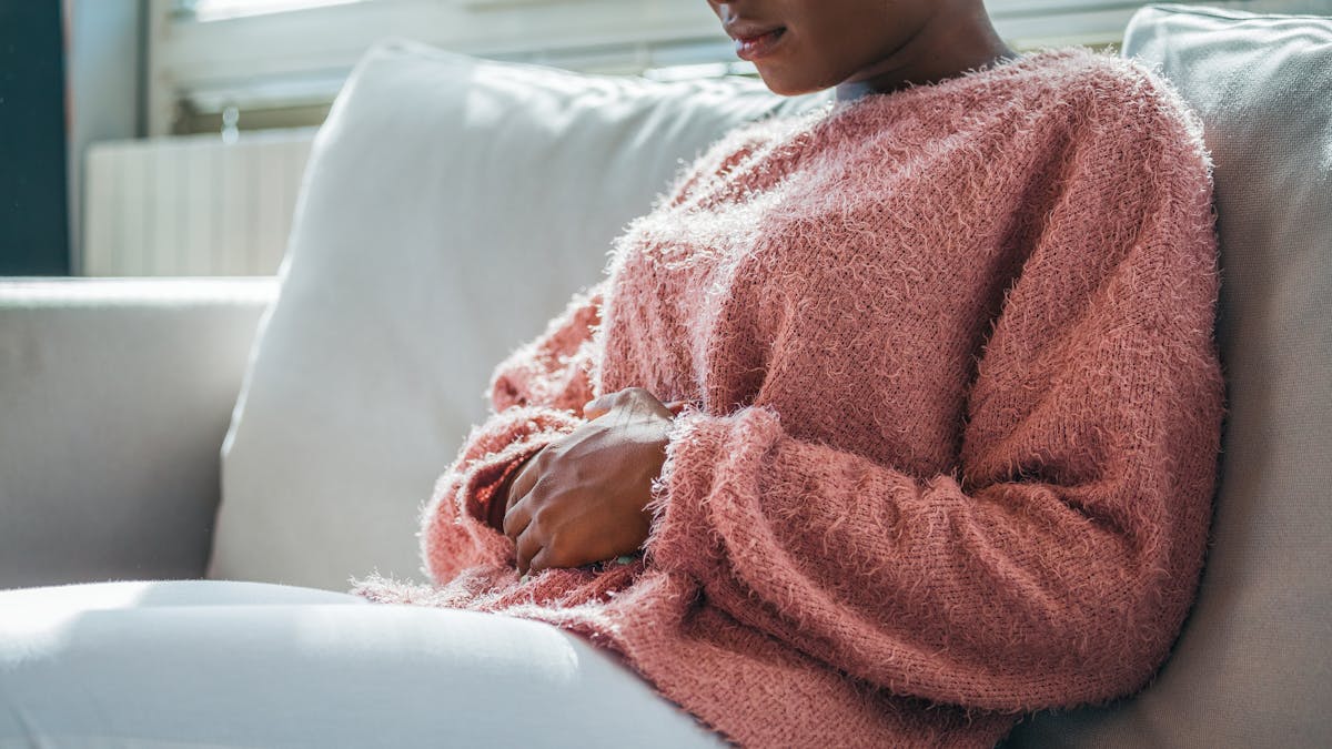 Woman in painful expression holding hands against belly suffering menstrual period pain, lying sad on home bed, having tummy cramp in female health concept