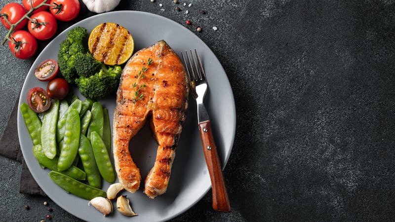 Tasty and healthy salmon steak with green peas, broccoli and tomatoes on a gray plate. Diet food on a dark background with copy space. Top view. Flat lay